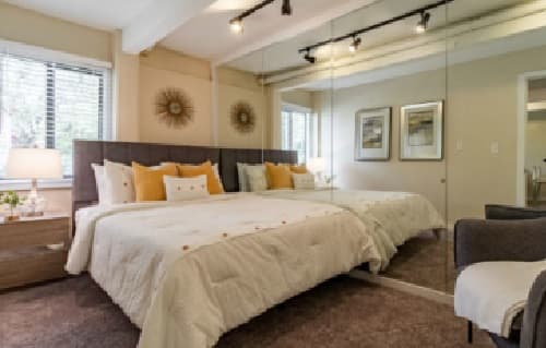 A modern bedroom at Buffalo Canyon Apartments Boulder features a large bed with white and yellow pillows, mirrored closet doors, a bedside table with a lamp, wall art, and a window letting in natural light.