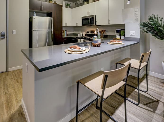 Modern kitchen with a gray countertop island, two wooden barstools, stainless steel appliances, and white and dark brown cabinets. Two plates with food are set on the island. A potted plant is visible on the right.