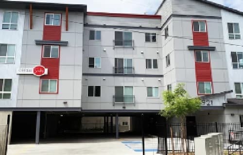 Modern four-story apartment building with white and gray exterior, red accents, several windows, balconies, a gated entry, a small tree, and covered parking on the ground level.