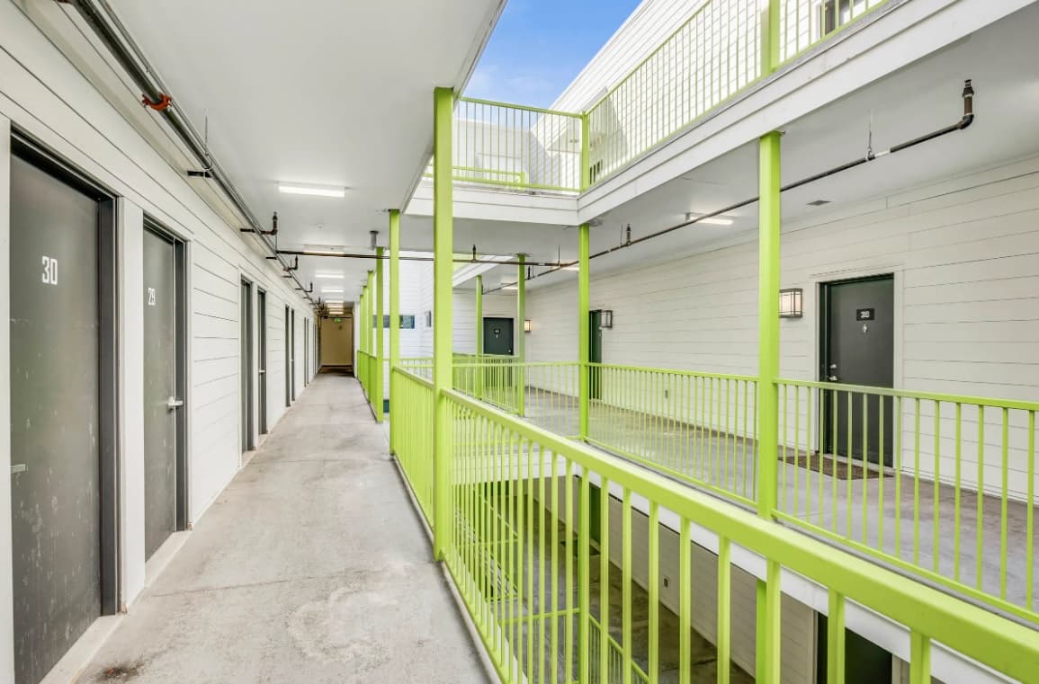 Second-floor walkway at Depot Square Apartments Boulder, featuring lime green railings, white walls, and numbered dark gray doors. The open corridor overlooks the level below and is exposed to the sky on one side.
