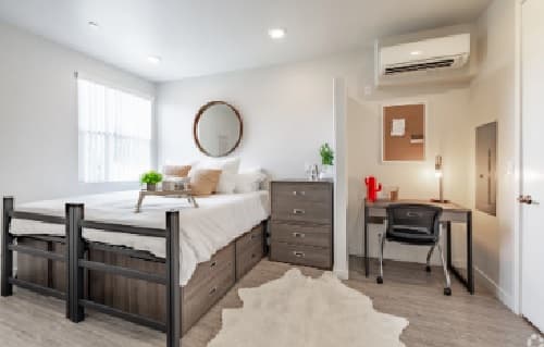 A modern, tidy bedroom at Element by Stuho features a bed with storage drawers, a dresser, a desk with chair, a corkboard, and a round mirror above the bed. Light-colored walls and a soft rug create a cozy feel.