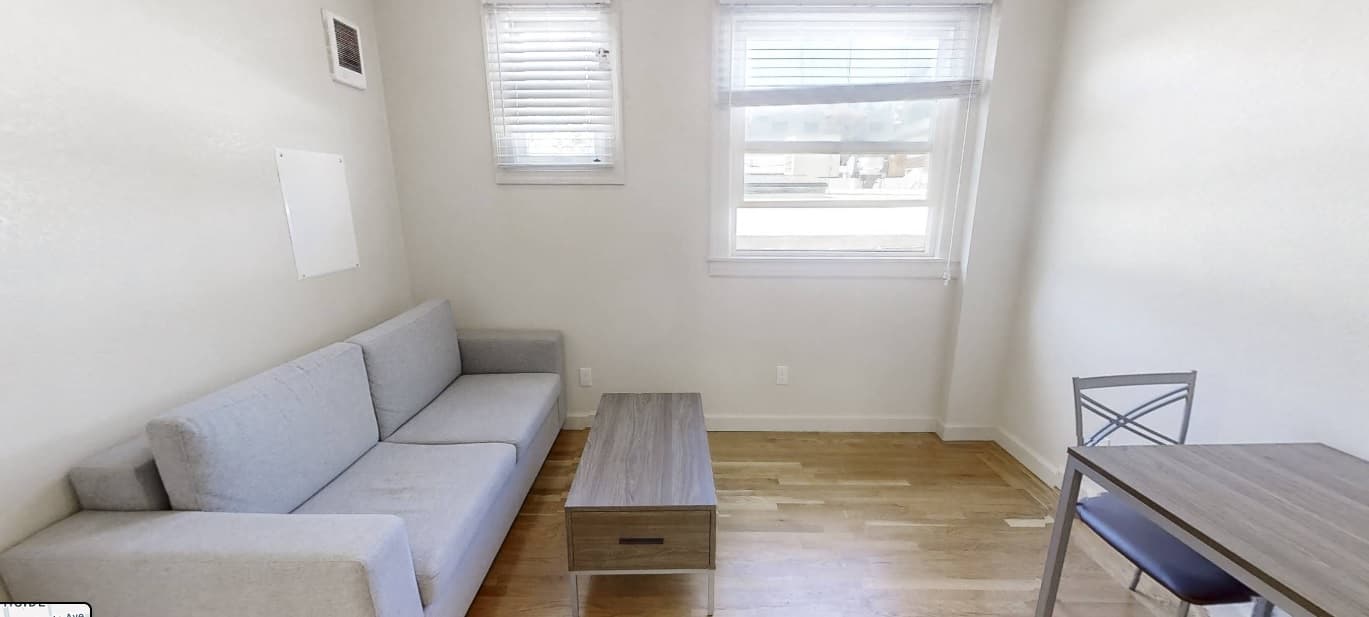 A small, bright room with light-colored walls, a gray sofa, a wooden coffee table, a metal chair, and a small table. Two windows let in natural light, and the floor is light wood.