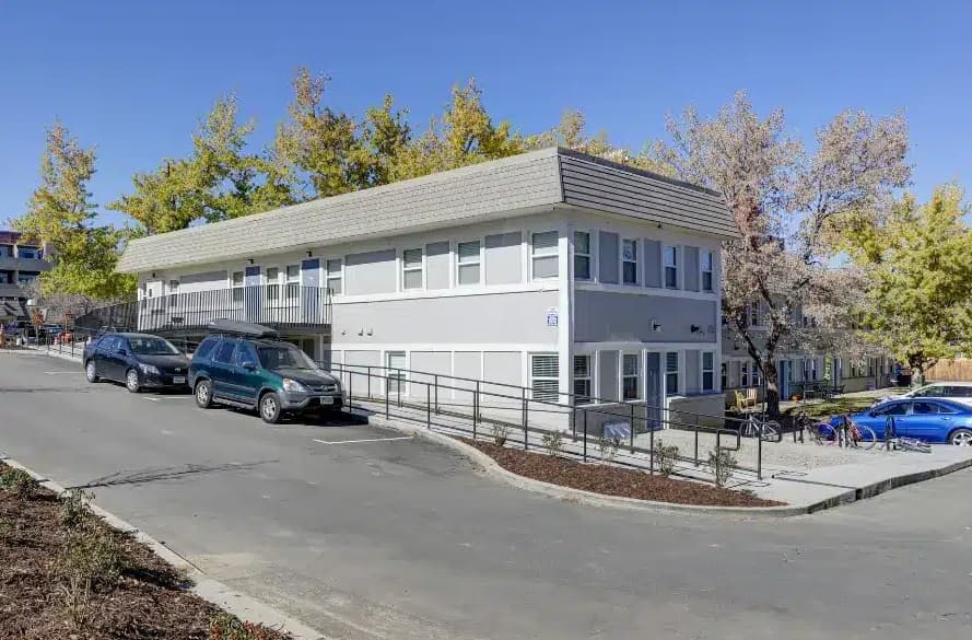 East Village Flats Boulder is a two-story, modern light gray building with large windows and a sloped roof, surrounded by parked cars and trees with yellow leaves on a sunny day; a ramp leads to the entrance.