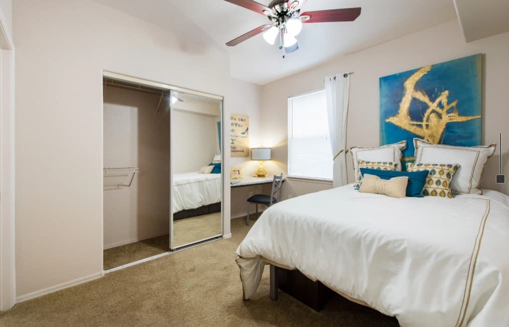 Bright bedroom at Gateway at Tempe with beige carpet, a bed with white and gold bedding, a large abstract painting above the bed, a small desk by the window, and a mirrored closet door reflecting the room.