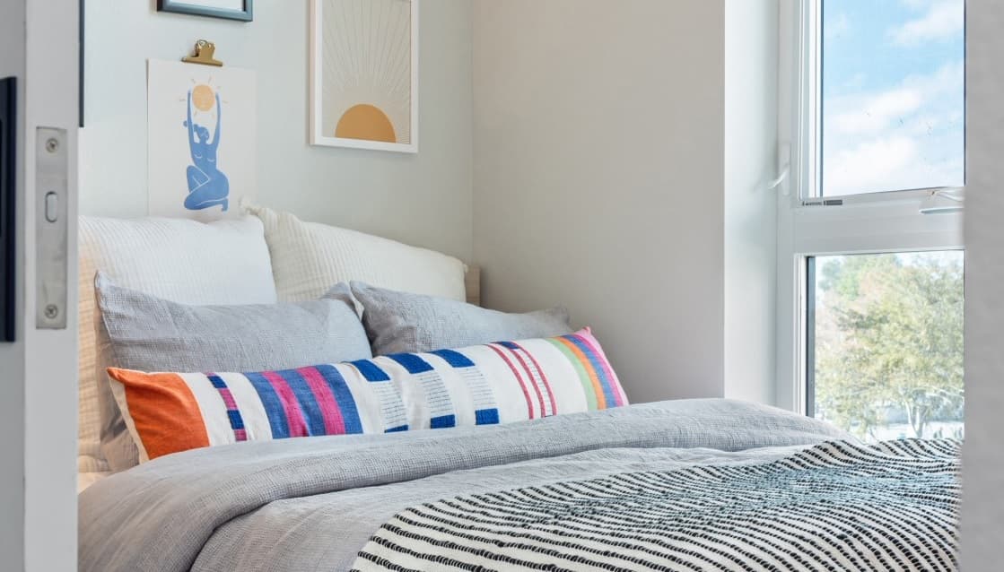 A cozy bedroom with a neatly made bed featuring gray and white bedding, a colorful patterned pillow, and framed art on the wall. Natural light streams in through a large window, echoing the bright ambiance of Hub Los Angeles.