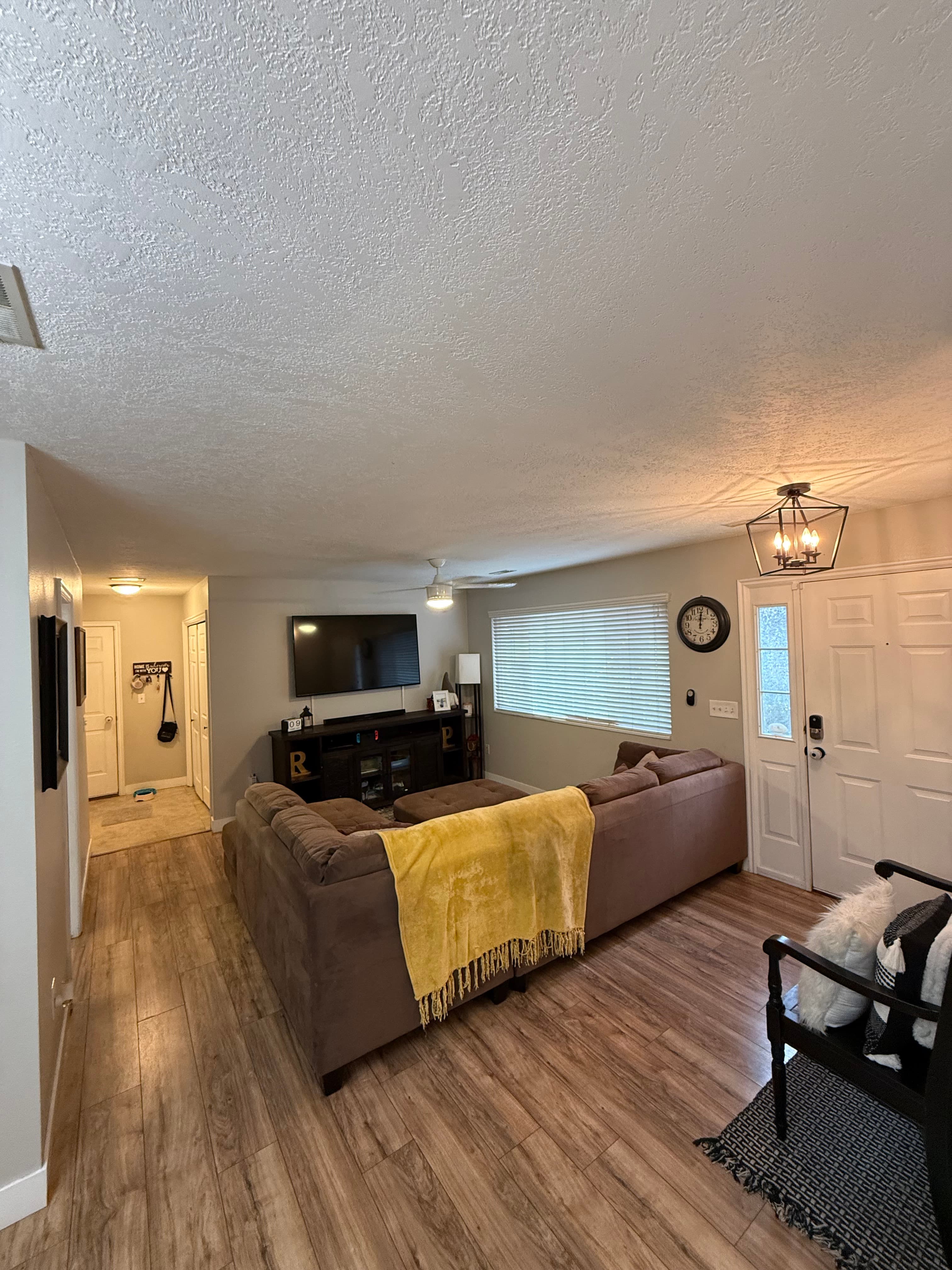 A cozy apartment living room at 282 W 960 S features a brown sectional sofa with a yellow blanket, a TV on a cabinet, wall clock, wooden floors, and a front door beside a window with closed blinds.