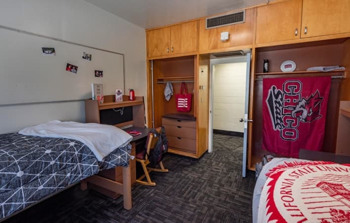 A neatly arranged college dorm room with two beds, open wooden closets, a Chico State banner, a chair, and some personal items on the desks and walls. The floor is carpeted and the room looks bright and tidy.