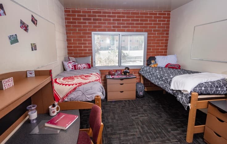 A college dorm room with two beds, each with different bedding, a window between them, two desks with chairs, and personal items like photos, books, and decorations on the desks and walls.