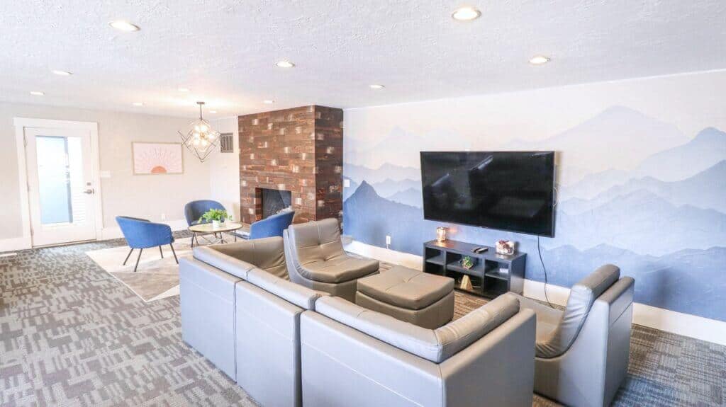 Modern living room at Liberty on Eighth Provo featuring a gray sectional sofa, blue accent chairs, wall-mounted TV, geometric carpet, wooden accent wall with fireplace, and a mountain-themed mural. Coffee table and decor complete the cozy space.