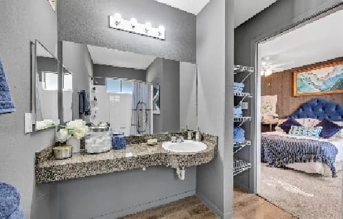 Modern bathroom with a granite countertop, sink, large mirror, and decorative items. Towels are neatly arranged on a shelf. A connected bedroom with a blue upholstered bed and colorful artwork is visible through an open doorway.