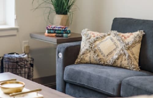 A cozy living room scene at Sierra Student Apartments features a gray sofa, a patterned decorative pillow, a side table with stacked books and a plant, and a wire basket on the floor nearby.