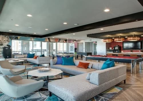 Modern lounge area with large windows, grey sectional sofas, colorful pillows, and a geometric rug. Pool tables and pendant lights in the background give this Saga Reno space a stylish, open entertainment vibe—perfect for relaxing or hosting friends.