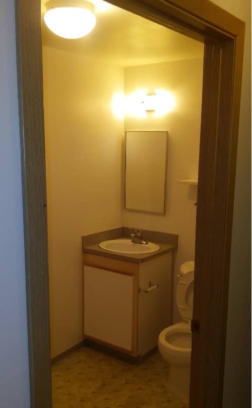 A small bathroom at Spencer View Apartments with yellow lighting features a sink with a mirror above, a cabinet below, and a toilet to the right. The floor has a light patterned design while the walls remain plain.