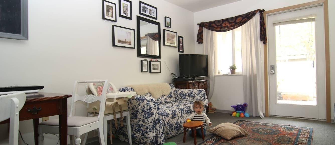 A living room at Spencer View Apartments features a floral sofa, a small child playing with toys on the floor, wall art above the couch, a TV in the corner, a curtained window, and a glass door leading outside.