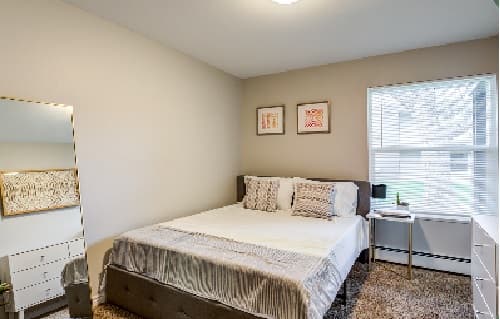 A neatly made bed with light bedding in a bright, minimal bedroom at The Social West® Fort Collins; a window with blinds, a white desk and chair, a tall mirror, and wall art are also visible.