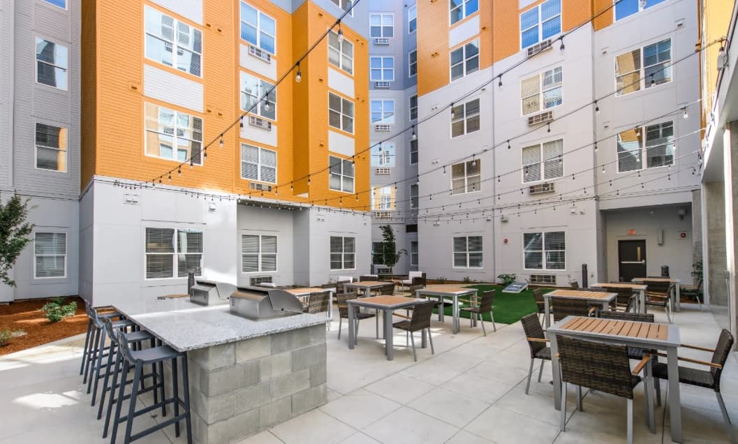 Modern Sierra Student Apartments courtyard with string lights, outdoor tables and chairs, a built-in grill, and a bar counter. Surrounded by multi-story buildings with orange and white exteriors and numerous windows.