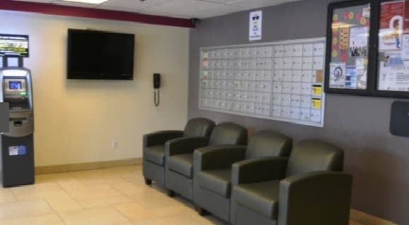 Four dark green chairs are lined up against a wall with mailboxes in a waiting area. There is an ATM, a wall-mounted TV, a phone, and a bulletin board with flyers. The floor is tiled.