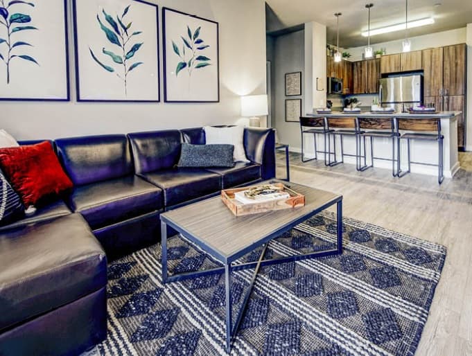 Modern living room at U Club on 28th Boulder with a dark leather sectional sofa, patterned rug, and wooden coffee table. Three botanical prints hang on the wall. The space flows into a kitchen with wood cabinets and a breakfast bar.