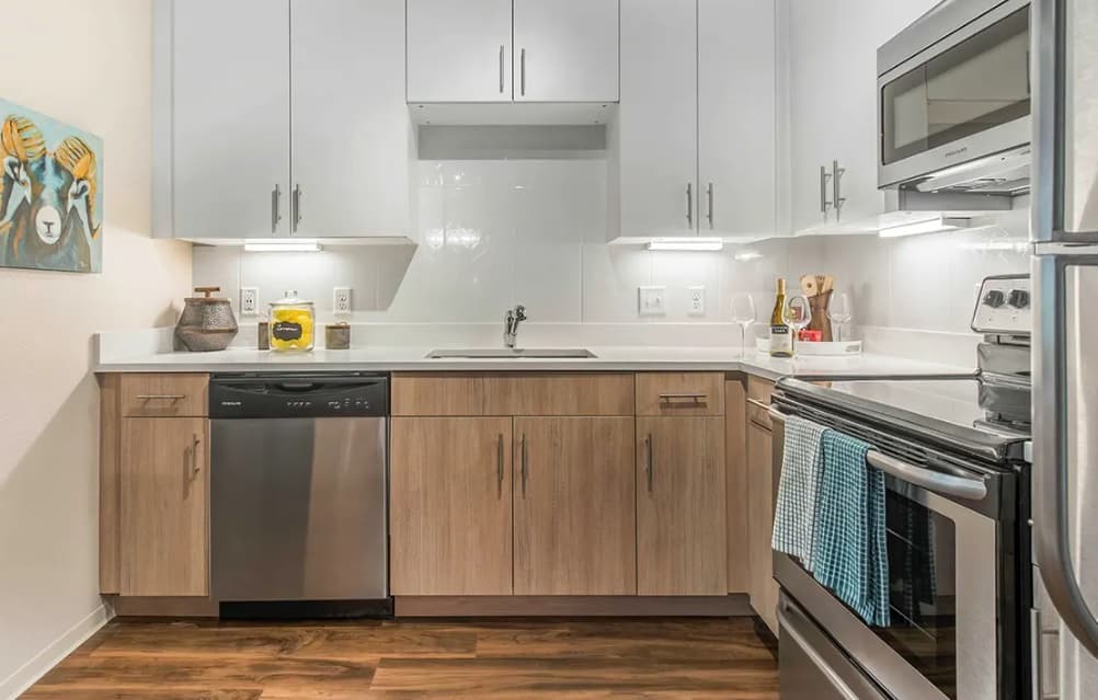Modern kitchen with light wood lower cabinets, white uppers, and stainless steel appliances brings an uncommon Fort Collins charm. A blue towel hangs on the oven door, and a quirky animal painting with hats is displayed on the left wall.