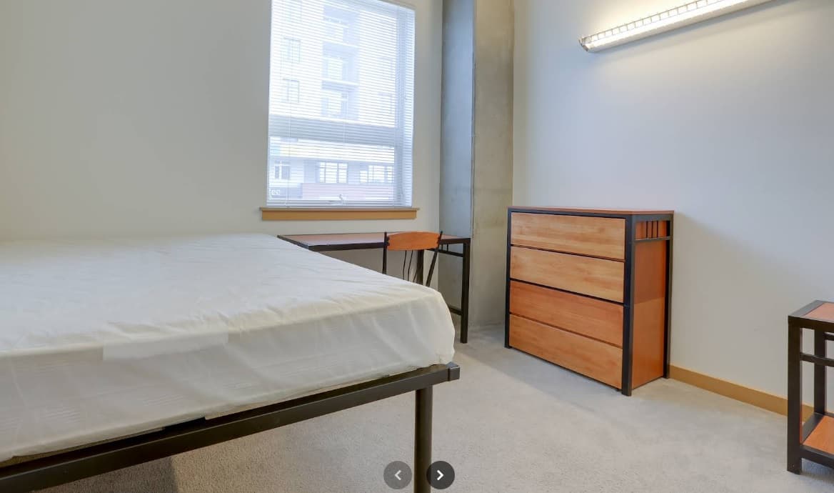 A minimalist University Lofts bedroom featuring a bed, wooden desk and chair by the window, a three-drawer wooden dresser, and beige carpeting. Natural light streams in through window blinds.