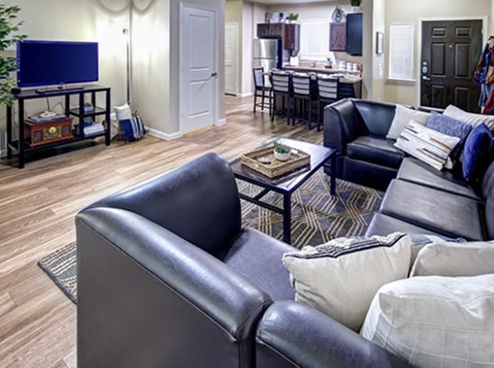 Modern living room at U Club Townhomes with dark leather sofas, coffee table, and TV on a stand. An open kitchen with bar stools is visible in the background—ideal student housing near Marion Pugh, featuring wooden floors and neutral walls.