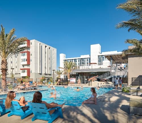 People relax and swim in an outdoor pool surrounded by lounge chairs and palm trees, with modern South Tower apartment buildings and a clear blue sky in the background at South Campus Plaza campus housing.