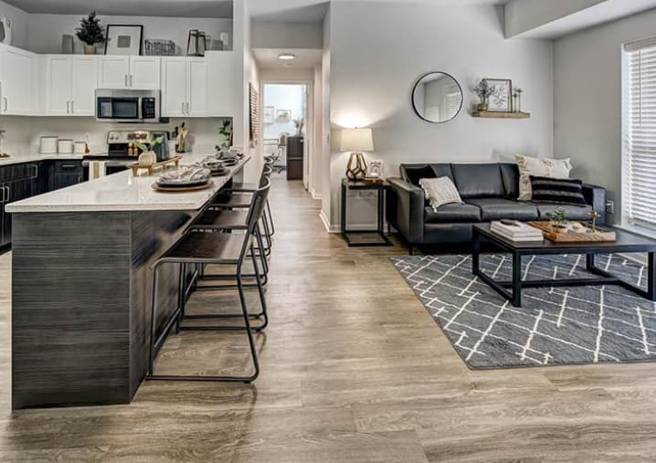 Modern open-concept living space at University Village at Boulder Creek Boulder with a kitchen island and bar stools, black sofa, coffee table, geometric rug, wall mirror, and large windows letting in natural light.
