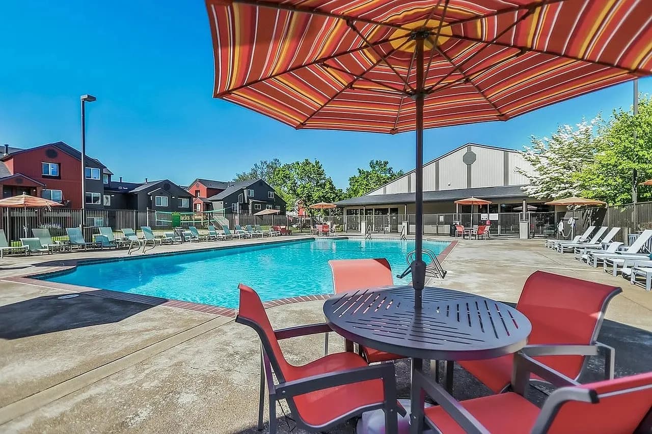 Aerial view of Ducks Village apartment complex with a swimming pool, hot tub, lounge chairs, sand volleyball court, parking lot, and autumn trees. Nearby buildings and cars are also visible.