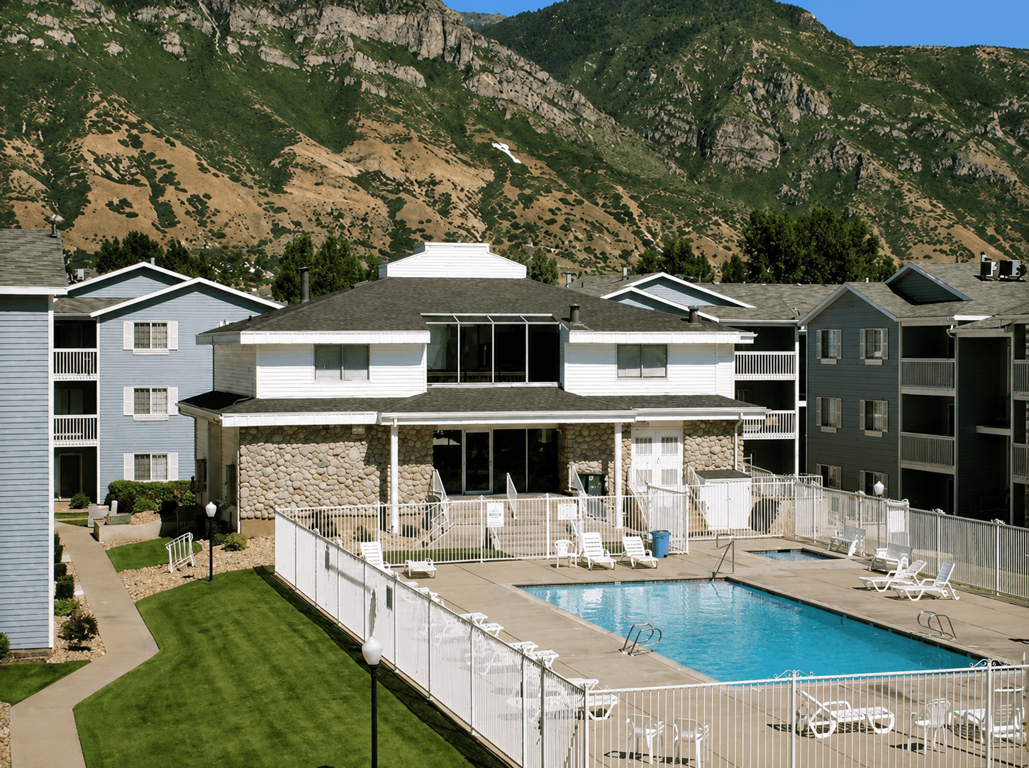 An outdoor swimming pool at Carriage Cove Provo is surrounded by a white fence, nestled between light blue apartment buildings, with mountains in the background under a partly cloudy sky.