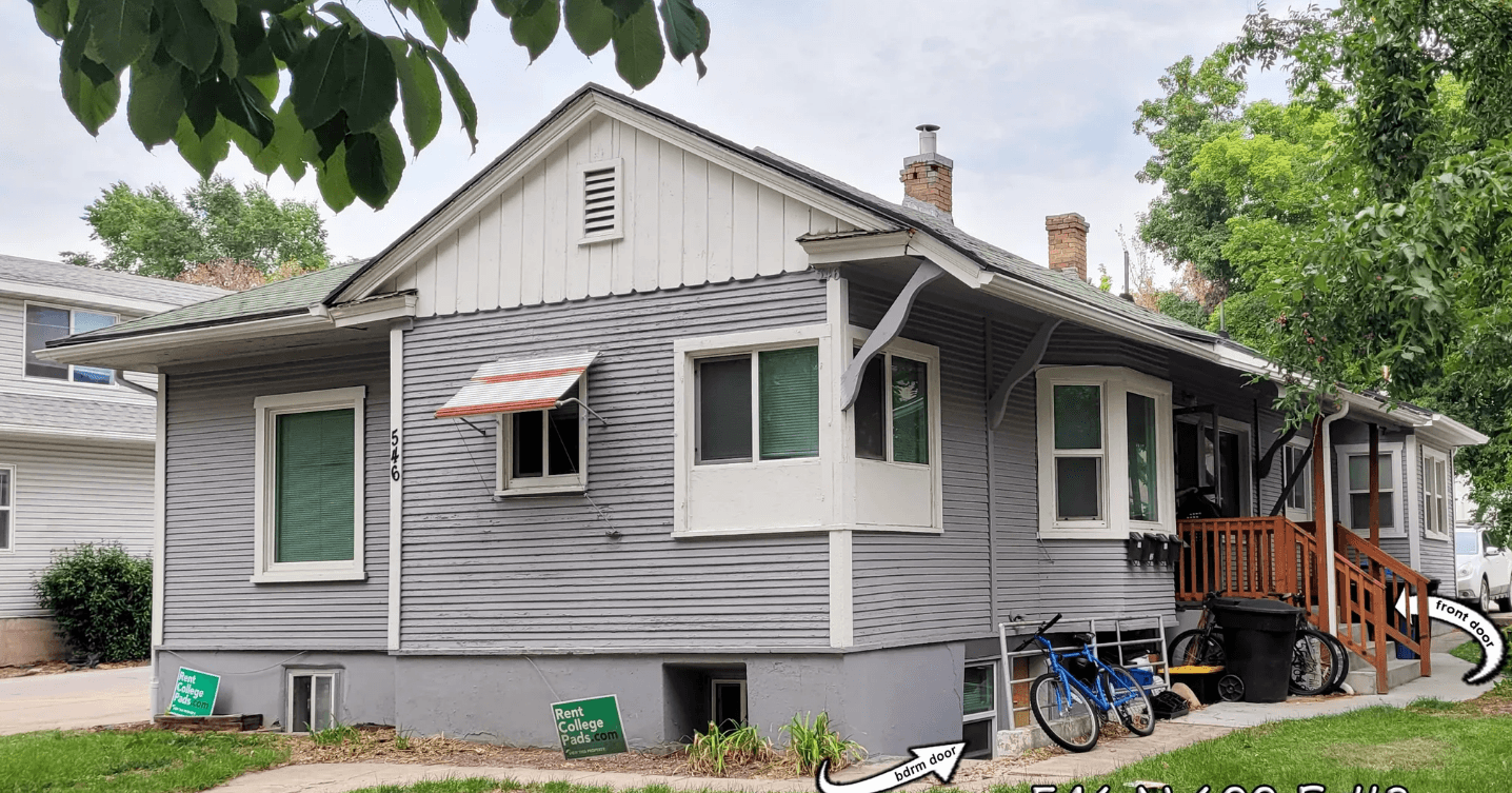 Gray single-story house with white trim and a small porch. Bicycles are next to the porch, and a white arrow points to a basement window labeled 546 N 600 E #3, part of Autumn Hills 546 Logan. Green lawn signs are visible in front.