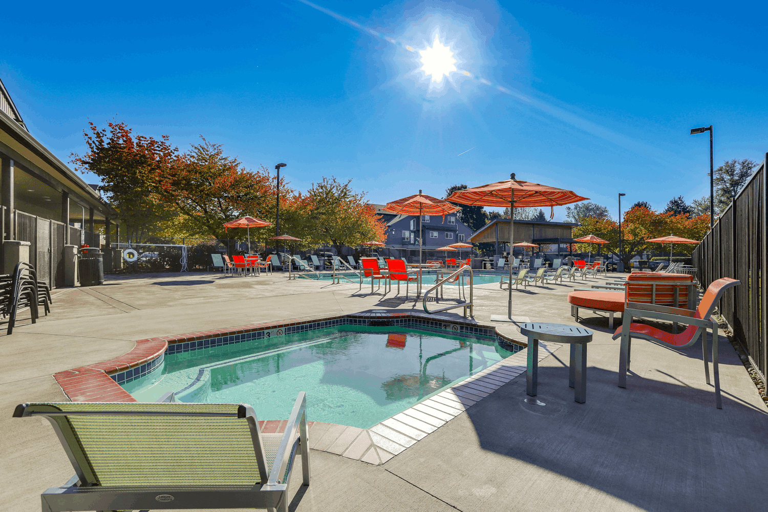 Aerial view of Ducks Village apartment complex with a swimming pool, hot tub, lounge chairs, sand volleyball court, parking lot, and autumn trees. Nearby buildings and cars are also visible.