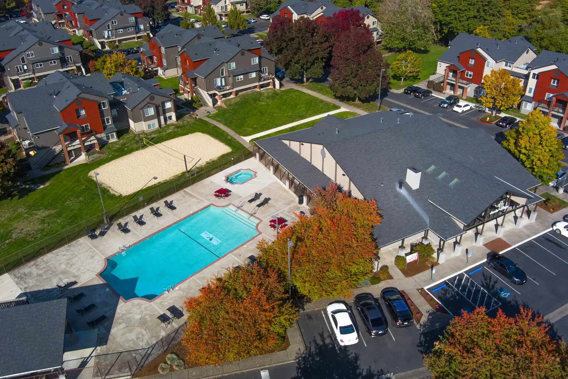 Aerial view of Ducks Village apartment complex with a swimming pool, hot tub, lounge chairs, sand volleyball court, parking lot, and autumn trees. Nearby buildings and cars are also visible.