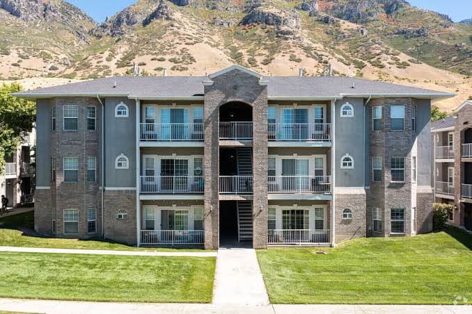 Gray Harris Condo buildings surrounded by green lawns and trees, with mountains and a partly cloudy blue sky in the background.