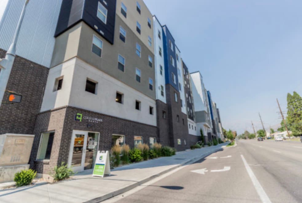 The photo shows the exterior of CollegePlace Provo, a modern multi-story building featuring a sign above the entrance. The facade combines sleek dark brick with light-colored concrete for a contemporary look.