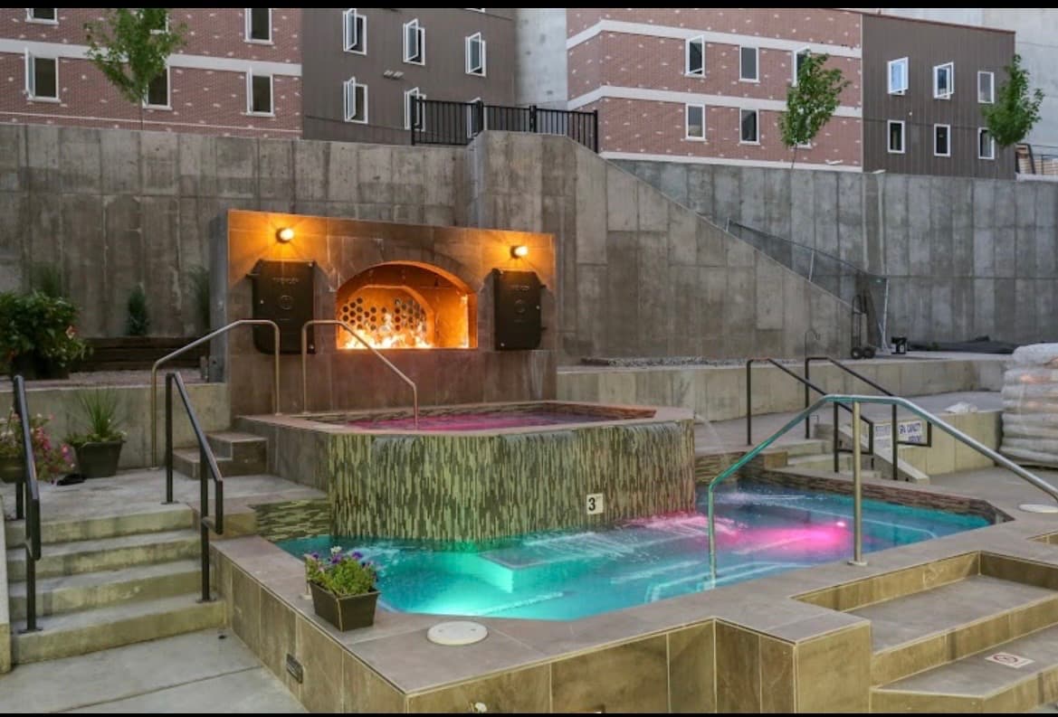 A modern, multi-story apartment building—The Factory Student Housing Logan—features brown and black exterior panels, large windows, and a landscaped area with orange flowers and greenery along the sidewalk under a clear blue sky.