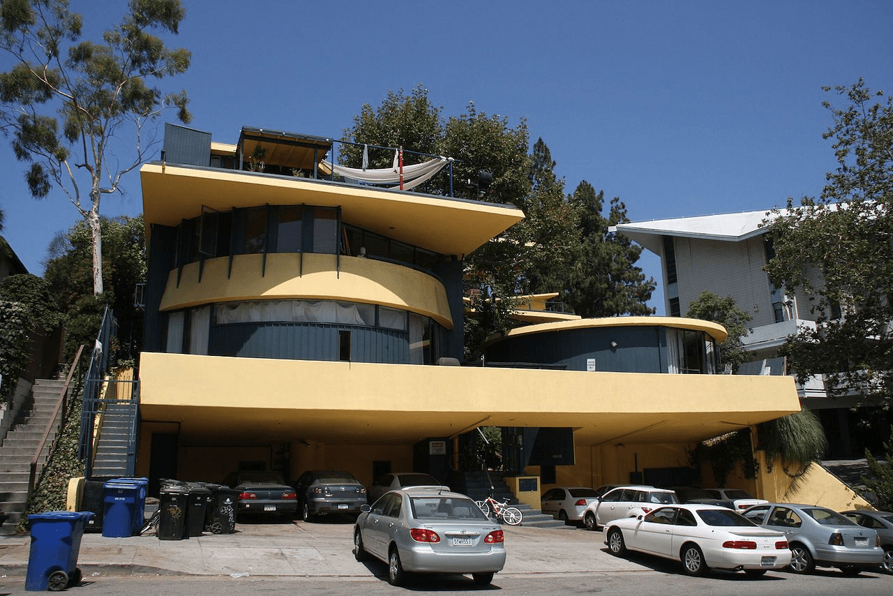 A modern, multi-story apartment building—The Mark Los Angeles—features striped gray metal siding and many windows. A small landscaped area with grass and shrubs sits in front, along with a utility box on the sidewalk beneath a clear blue sky.
