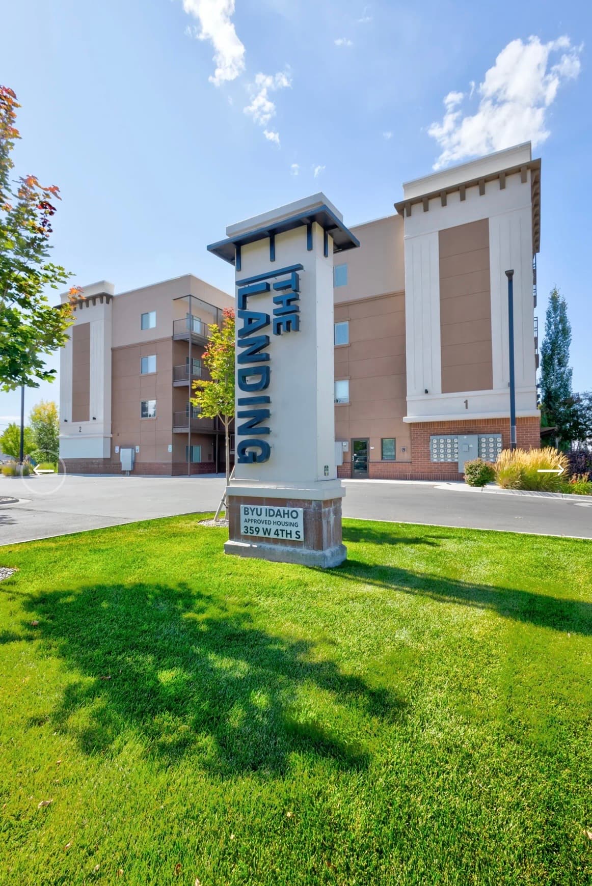 A modern, four-story apartment building with tan and white exterior walls stands behind a green lawn with a tall, vertical sign reading INDIGO near The Landing Rexburg driveway and parked cars.