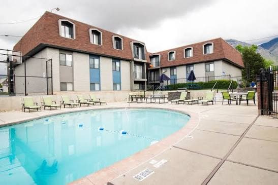 Liberty on Eighth Provo features a three-story apartment building with a brown mansard roof, white and blue-gray exterior walls, several windows, shrubs, a small tree, and a sidewalk in front.
