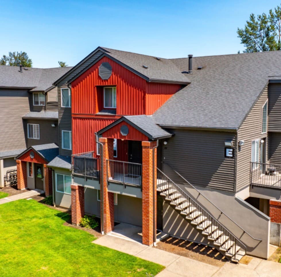 Aerial view of Ducks Village apartment complex with a swimming pool, hot tub, lounge chairs, sand volleyball court, parking lot, and autumn trees. Nearby buildings and cars are also visible.