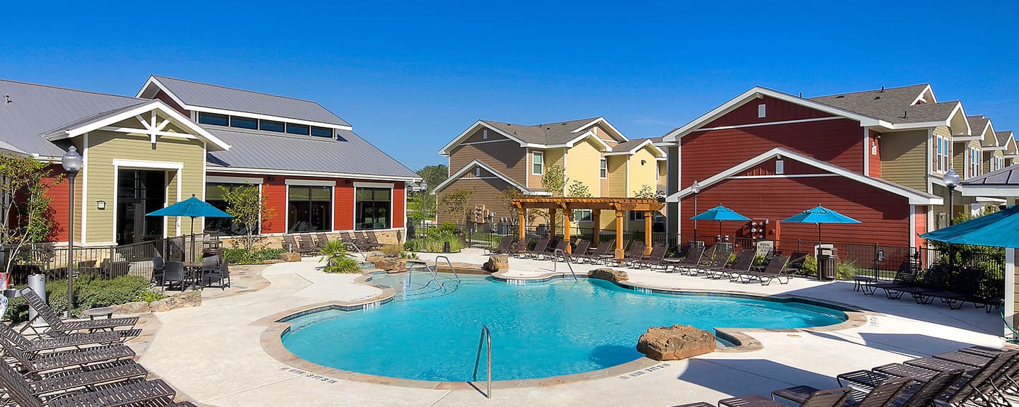 Row of modern, two-story U Club Townhomes with beige and red facades, white trim, small front porches, and empty parking spaces—ideal student housing near Marion Pugh under a clear blue sky.