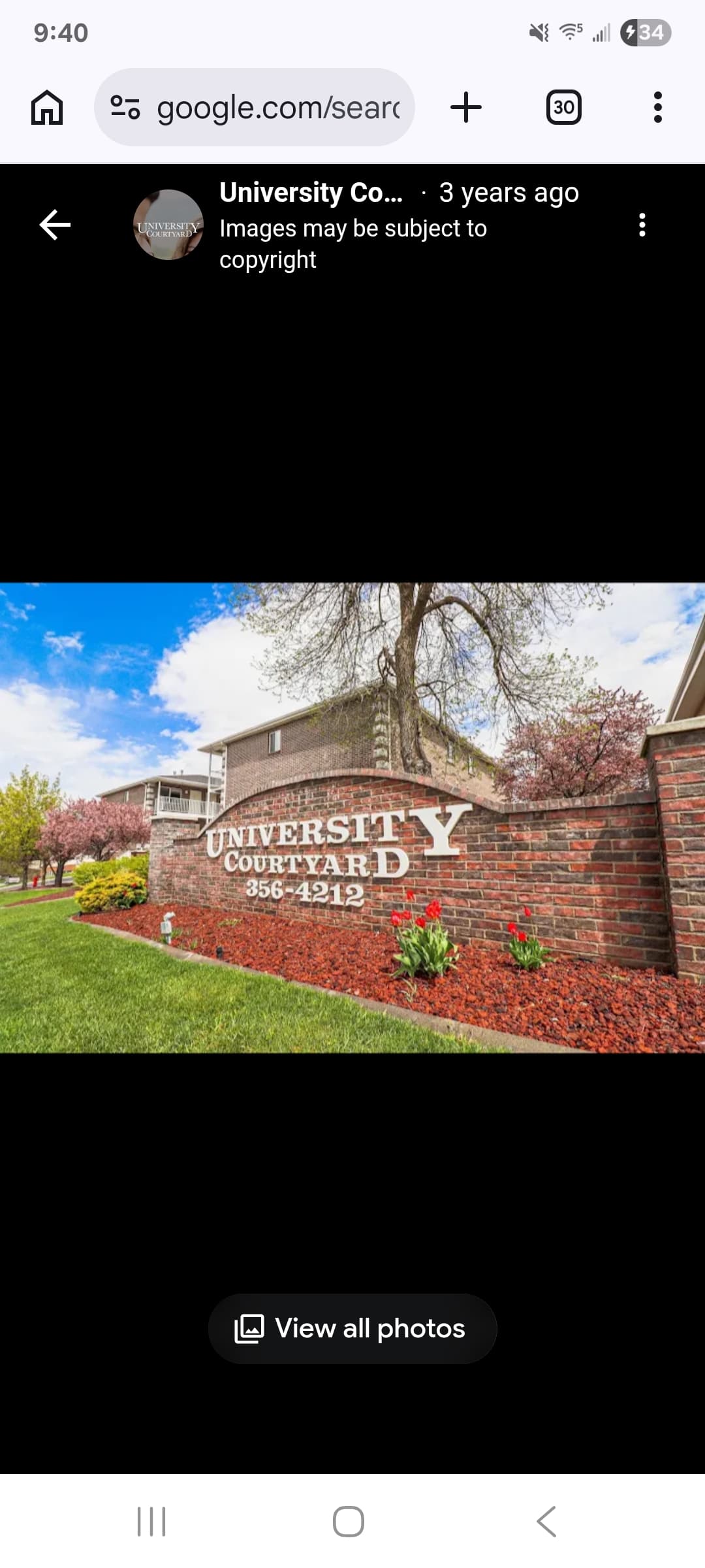 A three-story brick apartment building at University Courtyard Rexburg features exterior staircases and balconies on a sunny day. Several parked cars, bicycles near the entrance, and autumn-colored trees complete the scene.