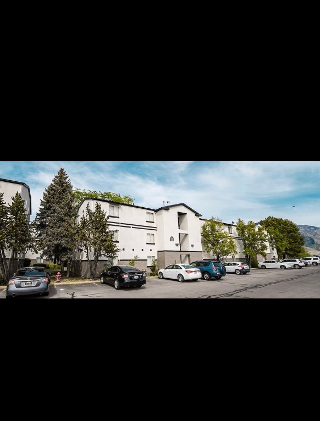 A three-story white apartment building with balconies and dark trim at Crestwood Provo, several parked cars in front, viewed from a parking lot on a cloudy day.