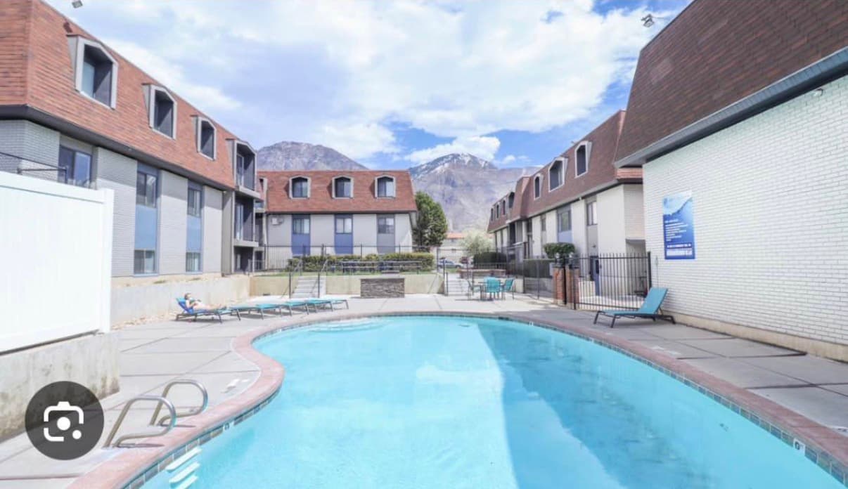 Liberty on Eighth Provo features a three-story apartment building with a brown mansard roof, white and blue-gray exterior walls, several windows, shrubs, a small tree, and a sidewalk in front.