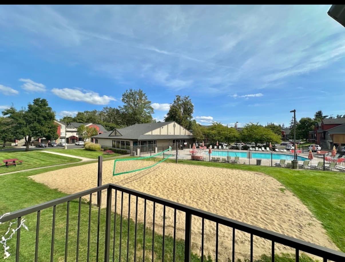 Aerial view of Ducks Village apartment complex with a swimming pool, hot tub, lounge chairs, sand volleyball court, parking lot, and autumn trees. Nearby buildings and cars are also visible.