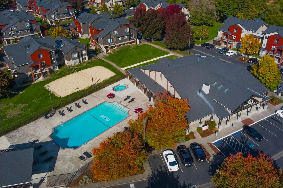 Aerial view of Ducks Village apartment complex with a swimming pool, hot tub, lounge chairs, sand volleyball court, parking lot, and autumn trees. Nearby buildings and cars are also visible.
