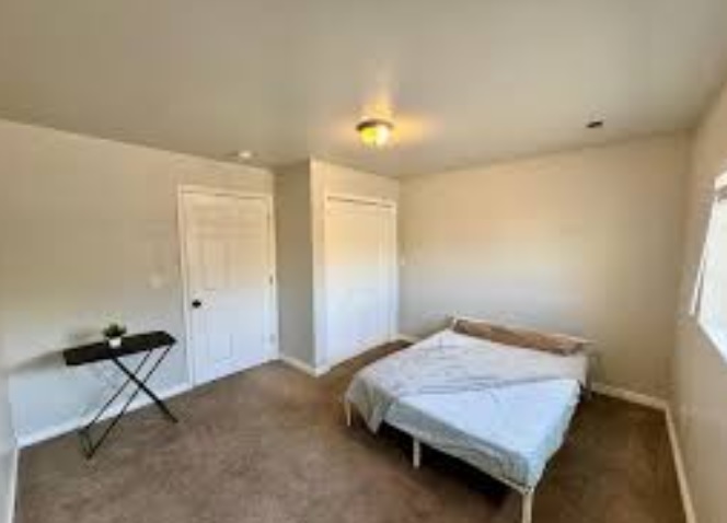 A simple Millcreek bedroom with beige carpet, white walls, a bed with white bedding, a small black folding table with a plant, two white closet doors, and a window letting in natural light.