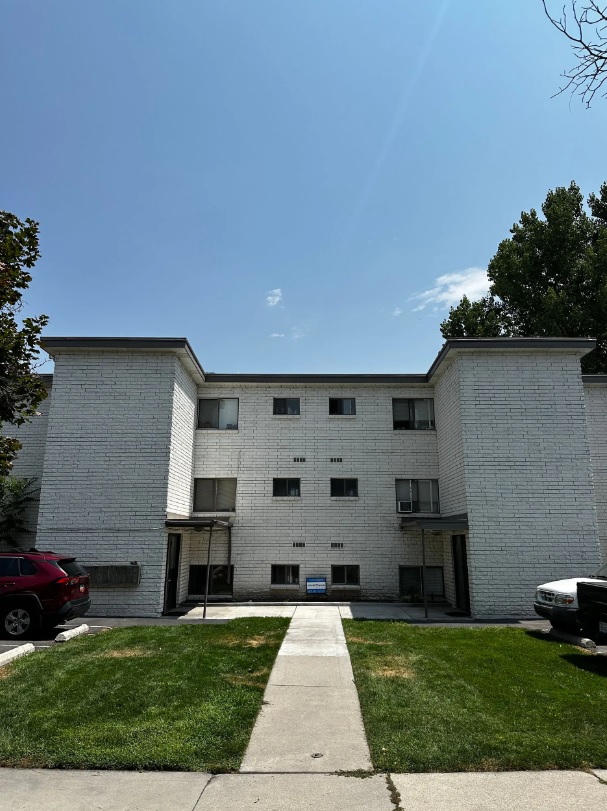 Autumn Apartments Provo is a three-story white brick building with small windows, two entrances, and a sidewalk leading to the doors. Cars line either side, and the lawn is green with some yellow patches.