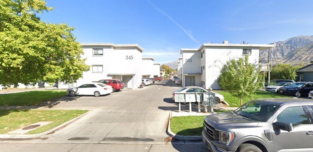 A wide driveway leads between two white three-story buildings labeled 345 and 363 at Lilac Place Apartments Provo. Several parked cars line the sides, with mountains visible in the background under a blue sky.