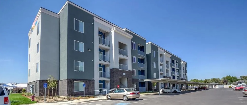 Modern three-story apartment building, The Boulevard Orem, features balconies, a gray and white exterior, and a front parking lot. Several cars are parked under a carport with visible handicap spaces, all beneath a clear blue sky.