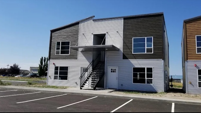 A modern two-story apartment building in Cantamere Village Rexburg features brown and white siding, large windows, an exterior staircase to the second floor, and an empty parking lot in front.